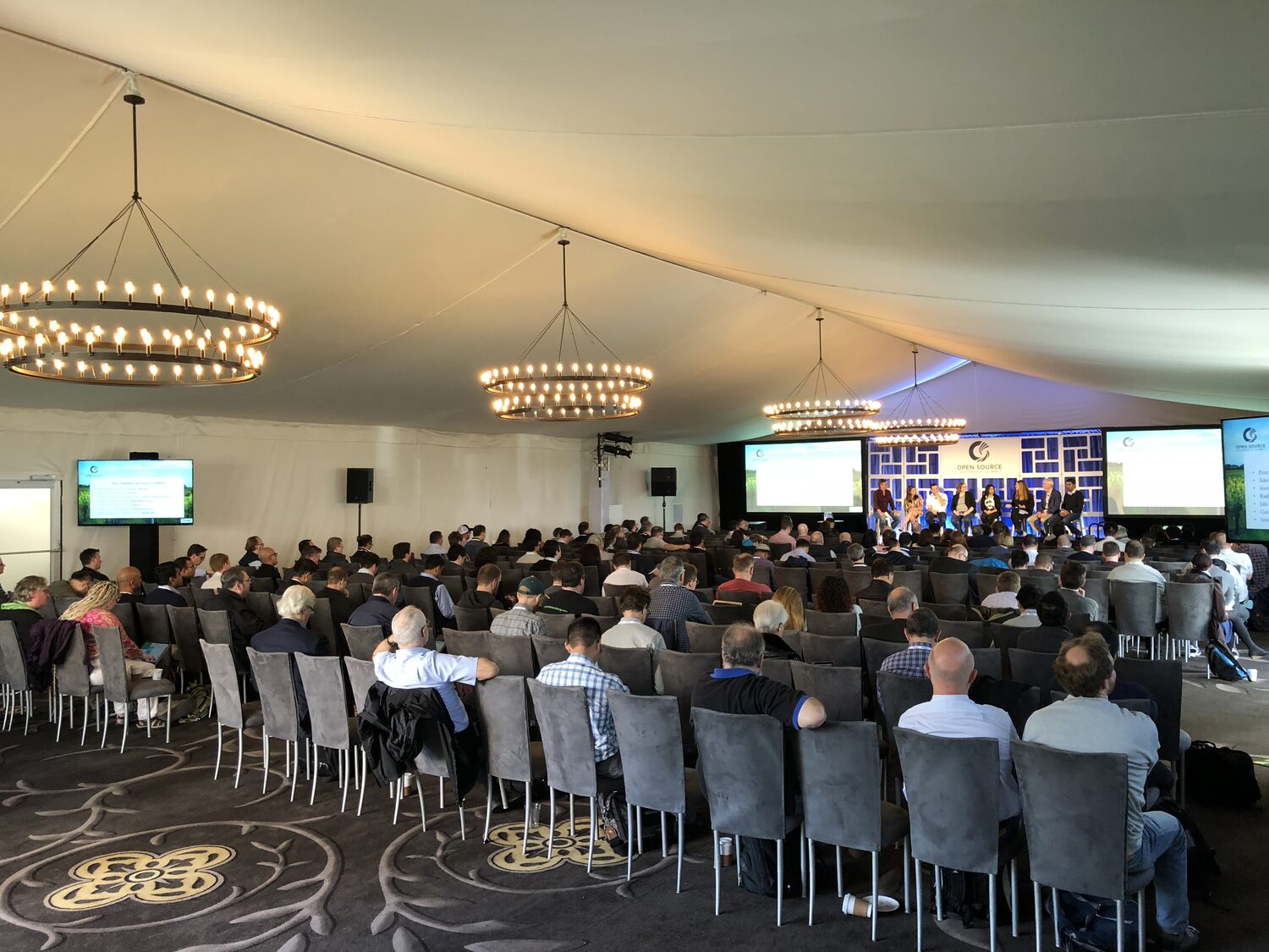 Delegates seated through the Pavilion marquee at the Open Source Leadership Summit, a seven-person panel on the lit stage under the Open Source logo, chandeliers hung from the canvas ceiling