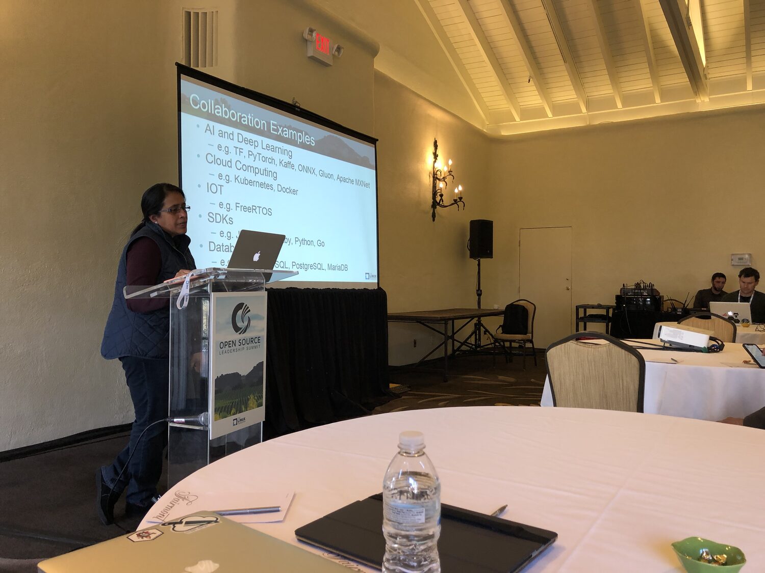 A speaker at the Open Source Leadership Summit lectern in a breakout room, slide behind reading Collaboration Examples with TensorFlow, PyTorch, ONNX, Gluon, Apache MXNet, Kubernetes and Docker listed