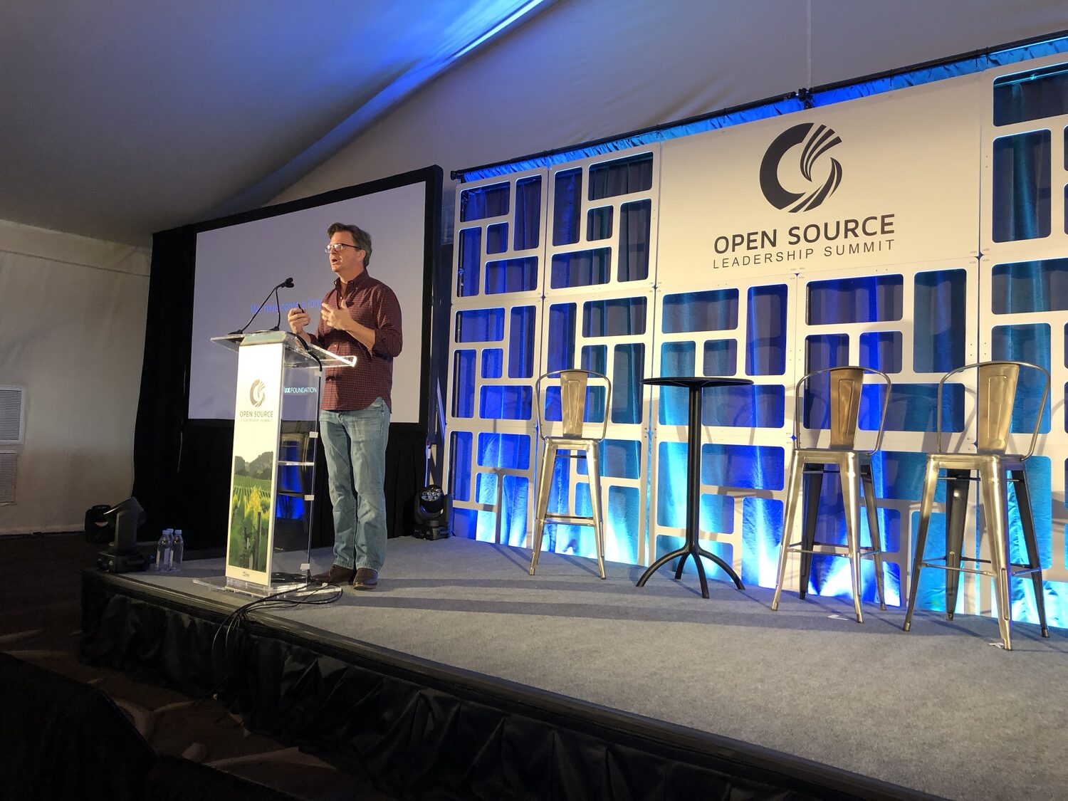 The summit keynote stage, Open Source Leadership Summit backdrop in blue and white, bar stools set for a panel and a speaker at the lectern