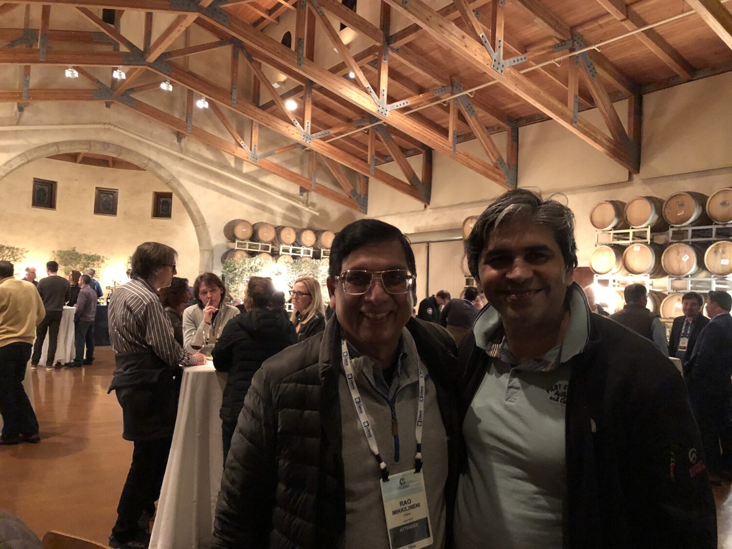 Tarry Singh with a fellow delegate in a winery barrel room at the summit evening reception, conference lanyards on, oak barrels racked along the wall behind