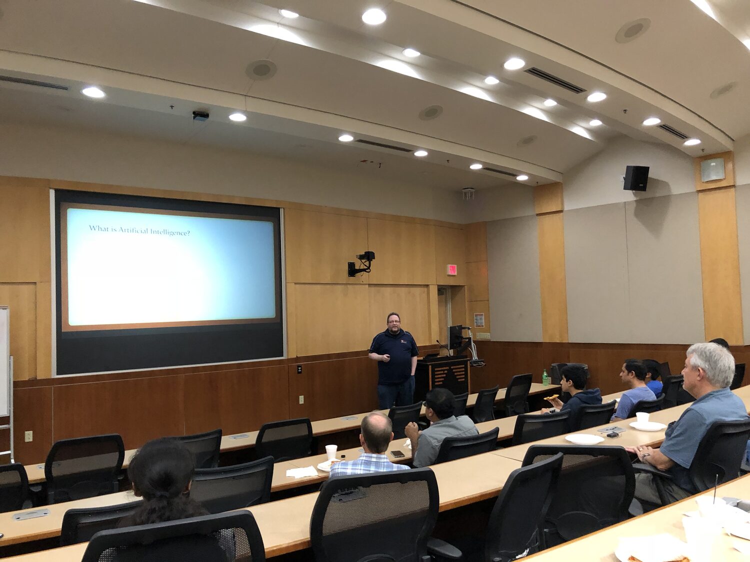 Evening session in a wood-panelled UT Dallas lecture room, a speaker at the podium and delegates at the desks