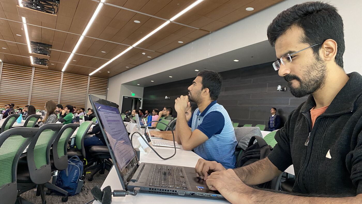 Two participants side by side at their laptops on the workshop floor, one in headphones, the hall full behind them.