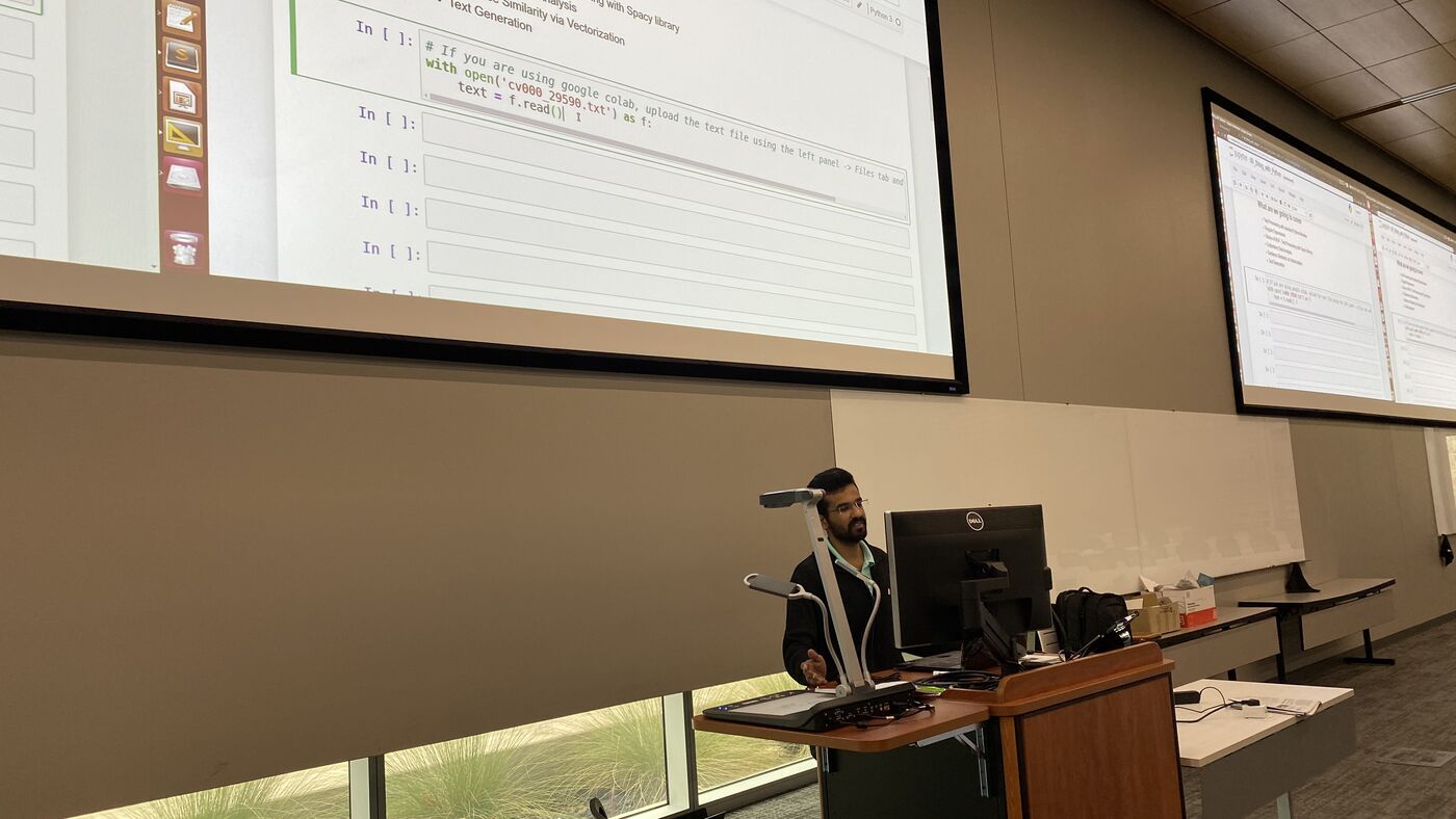A participant presenting from the lectern, screens lit behind.